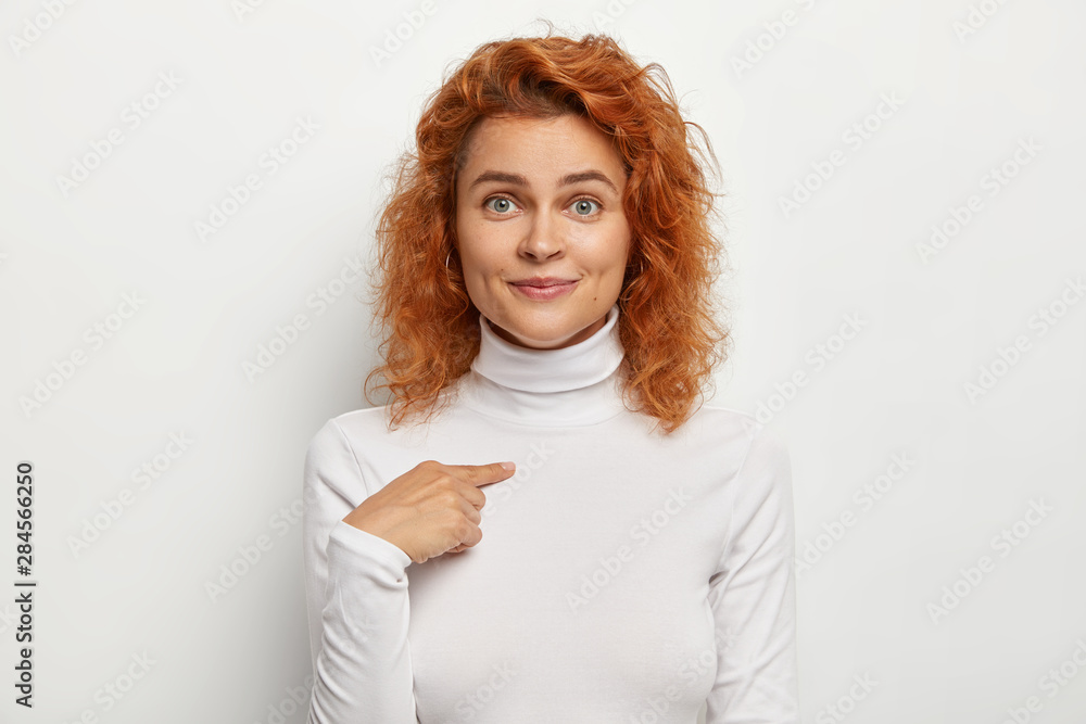 Pleasant looking young female points at herself, asks who me, has charming smile, blue eyes, natural red wavy hair, wears white turtleneck, poses against studio background. Do you choose me? - obrazy, fototapety, plakaty 