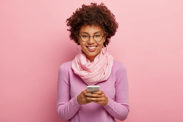 Cheerful smiling attractive female plays games on mobile phone application, sends text messages to friends, looks positively at camera, has curly Afro hairstyle, wears purple jumper and scarf on neck