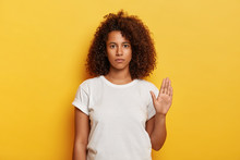 Stay Away From Me. Serious Attractive Girl With Curly Hair, Keeps Palm In Stop Gesture, Looks Confidently At Camera, Wears White T Shirt, Isolated On Yellow Background, Says Its Enough. Body Language