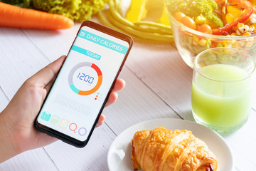 Wall Mural - Calories counting and food control concept. woman using Calorie counter application on her smartphone with salad , vegetable, juice and croissant on dining table