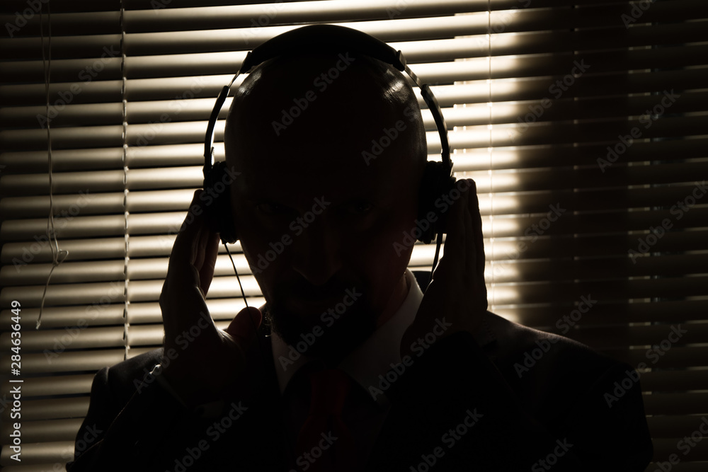 Bald man with headphones on the background of closed blinds, contour lighting, listening to music on old equipment - obrazy, fototapety, plakaty 