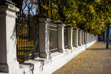 Sidewalk Along The White Stone Fence With Black Bars On The Background Of The Autumn Landscape. City Alley In Autumn.