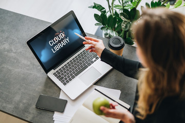 Woman is standing, pointing pencil on laptop computer with inscription on monitor- cloud library. Hipster girl working on pc.