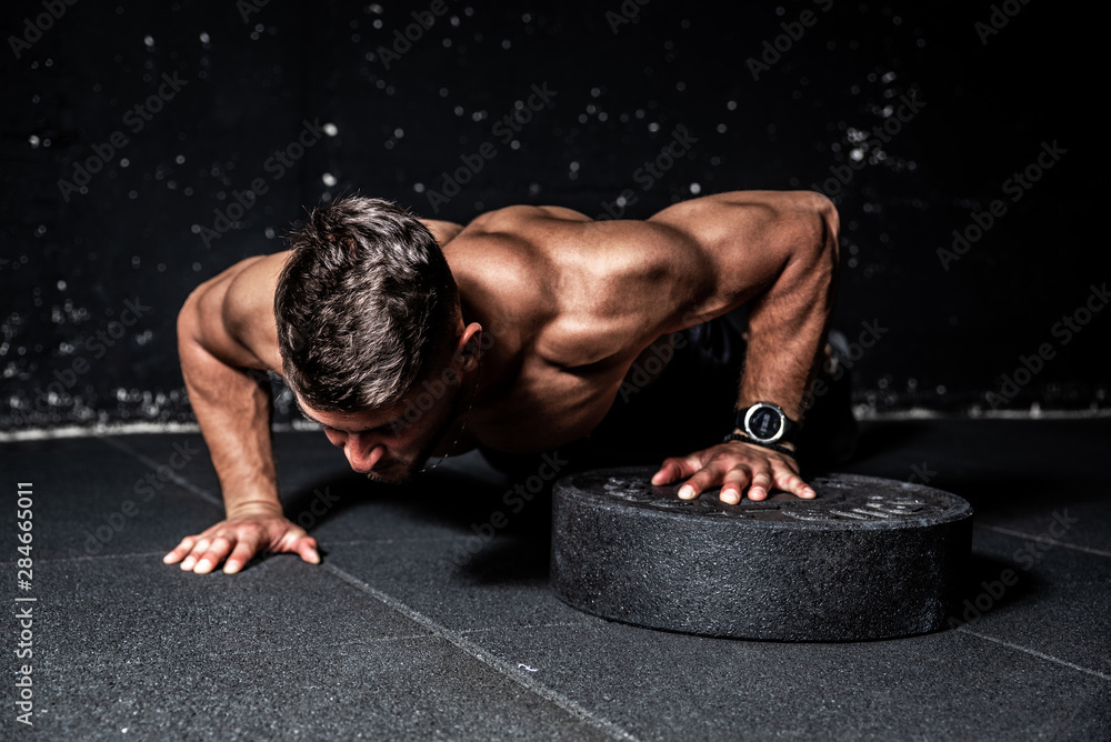 Young strong sweaty focused fit muscular man with big muscles performing push ups with one hand on the barbell weight plate for training hard core workout in the gym  - obrazy, fototapety, plakaty 