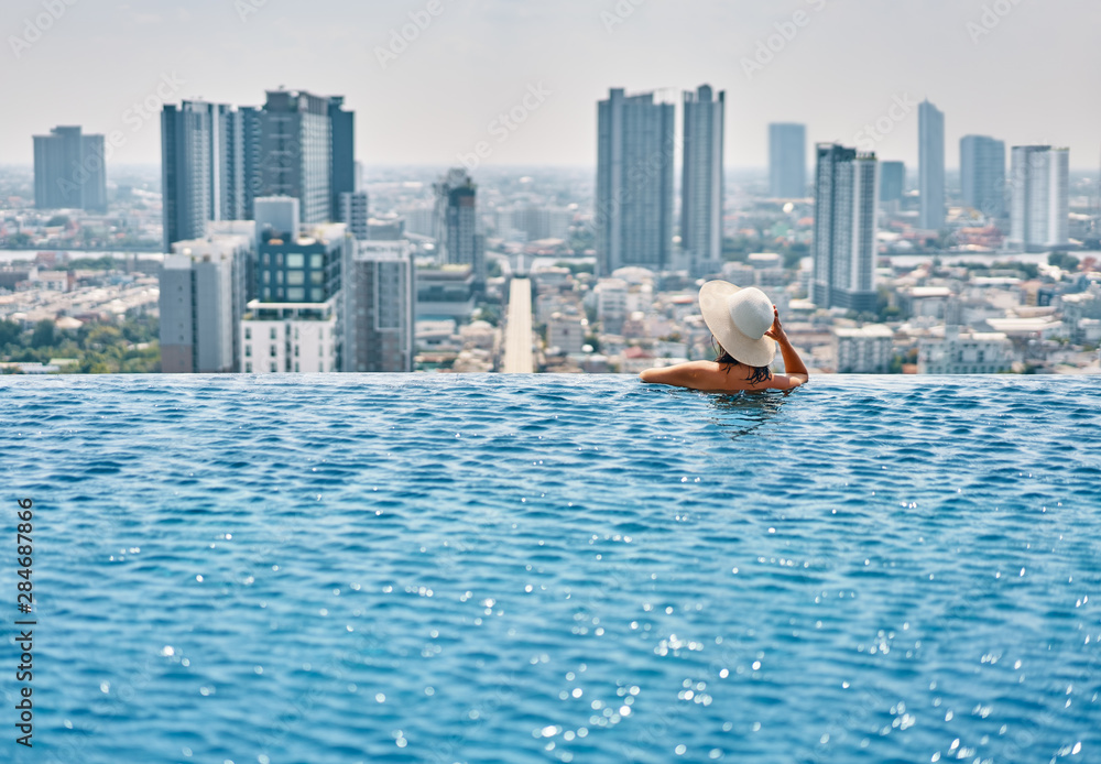 Back view of young woman in hat relaxing in swimming pool on the roof top of hotel and enjoy cityscape - obrazy, fototapety, plakaty 