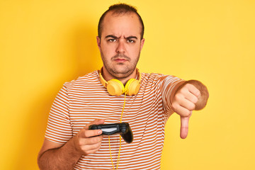 Canvas Print - Young gamer man playing video game using headphones over isolated yellow background with angry face, negative sign showing dislike with thumbs down, rejection concept