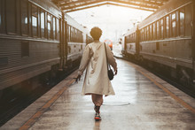 Rear View Of A Walking African Woman In A White Cloak On The Railway Station Before Her Trip Between Two Highway Trains Waiting For Departure On The Platform Indoors Of A Railroad Depot