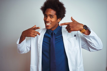 Poster - Young african american doctor man wearing coat standing over isolated white background smiling cheerful showing and pointing with fingers teeth and mouth. Dental health concept.