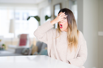 Canvas Print - Young beautiful woman wearing winter sweater at home peeking in shock covering face and eyes with hand, looking through fingers with embarrassed expression.