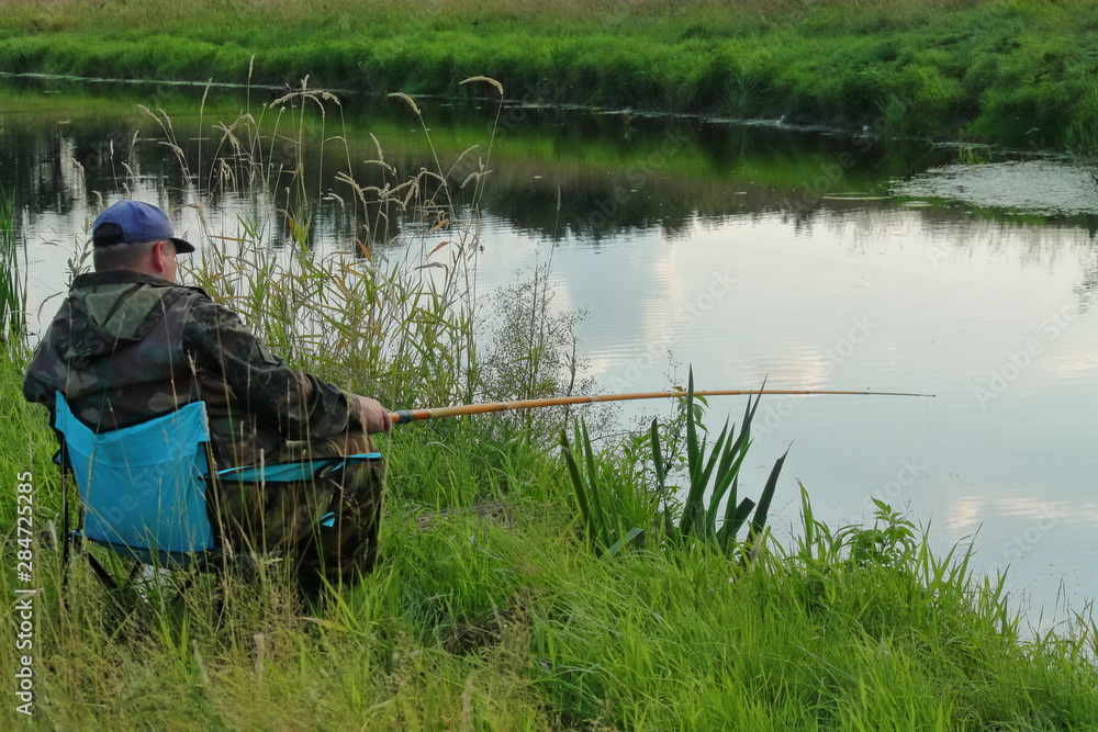 fisherman sitting in a chair and fishing. a man on the Bank of the river fishing rod. the fisherman enjoys fishing. man resting in nature. natural food for humans. Stock Photo |