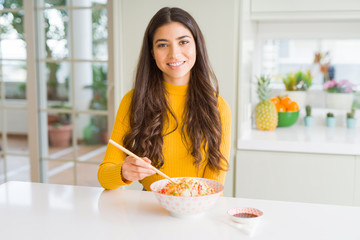 Poster - Young woman eating a bowl of Asian rice using chopsticks with a happy face standing and smiling with a confident smile showing teeth