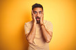 Young indian man wearing t-shirt standing over isolated yellow background afraid and shocked, surprise and amazed expression with hands on face