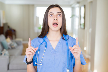 Poster - Beautiful young nurse woman wearing uniform and stethoscope at the clinic amazed and surprised looking up and pointing with fingers and raised arms.