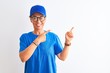 © Krakenimages.com - Senior deliverywoman wearing cap and glasses standing over isolated white background smiling and looking at the camera pointing with two hands and fingers to the side.