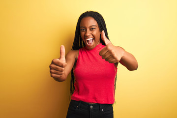 African american woman wearing red casual t-shirt standing over isolated yellow background approving doing positive gesture with hand, thumbs up smiling and happy for success. Winner gesture.