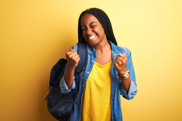 Poster - African american student woman wearing backpack standing over isolated yellow background very happy and excited doing winner gesture with arms raised, smiling and screaming for success. 