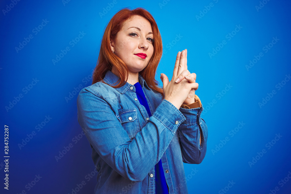Young beautiful redhead woman wearing denim shirt standing over blue isolated background Holding symbolic gun with hand gesture, playing killing shooting weapons, angry face
