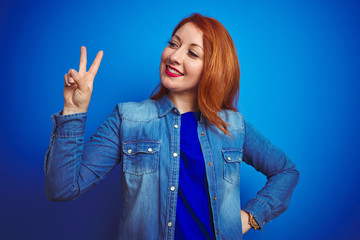 Poster - Young beautiful redhead woman wearing denim shirt standing over blue isolated background smiling looking to the camera showing fingers doing victory sign. Number two.