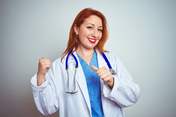 Sticker - Young redhead doctor woman using stethoscope over white isolated background Pointing to the back behind with hand and thumbs up, smiling confident