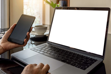 Wall Mural - Mockup image of businessperson hand  using mobile smart phone and touching laptop computer pad with blank white screen with cup of coffee on the desk at modern home office interior.  clipping path