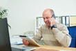 © DragonImages - Smiling mature entrepreneur reading information on digital tablet and talking on smartphone with clients when working at his office table
