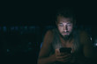 © Maridav - Dark night man looking at mobile phone screen texting late at night awake in bed insomnia or outside in city with skyline background. Serious looking guy depressed addicted to social media.