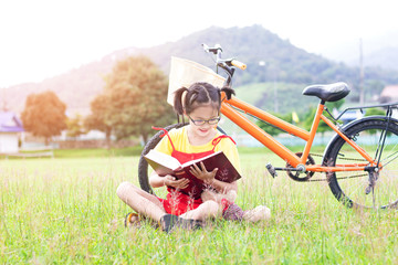 Poster - Cute asian student girl with glasses sitting on the grass reading a book in garden at summertime