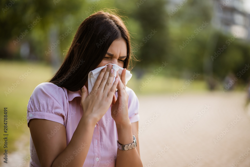 Woman with allergy symptom blowing nose in park. - obrazy, fototapety, plakaty 
