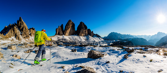 Fototapete - Mountaineer backcountry ski first snow walking up along a snowy ridge with skis. In background blue cloudy sky and shiny sun Tre Cime, Dolomites, Italy.