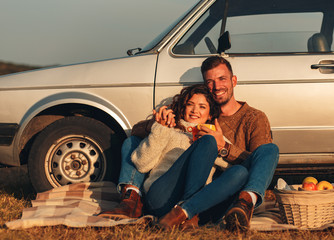 Wall Mural - Beautiful young couple enjoying picnic time on the sunset. They drinking tea and sitting in a meadow leaning on old fashioned car.