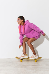 Canvas Print - Young african woman skater isolated over white wall background in bright pink sweatshirt on skateboard.