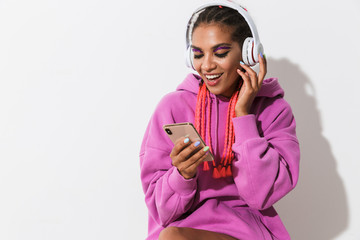 Poster - Pleased positive young african woman isolated over white wall background in bright pink sweatshirt using mobile phone listening music with headphones.