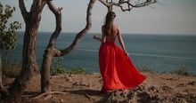 Back View Of Beautiful Woman In Red Dress Walking On The Sea Shore Cliff During Sunny Summer Day - Video In Slow Motion