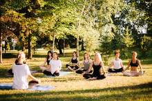 Group Of Young Women Are Meditating In Park On Summer Sunny Morning Under Guidance Of Instructor. Group Of Girl Outdoors Are Sitting In Lotus Pose On Yoga Mats On Green Grass With Eyes Closed