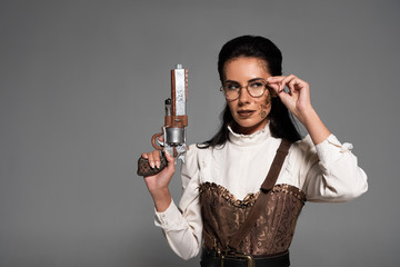 Wall Mural - pensive steampunk woman holding vintage pistol and touching glasses isolated on grey