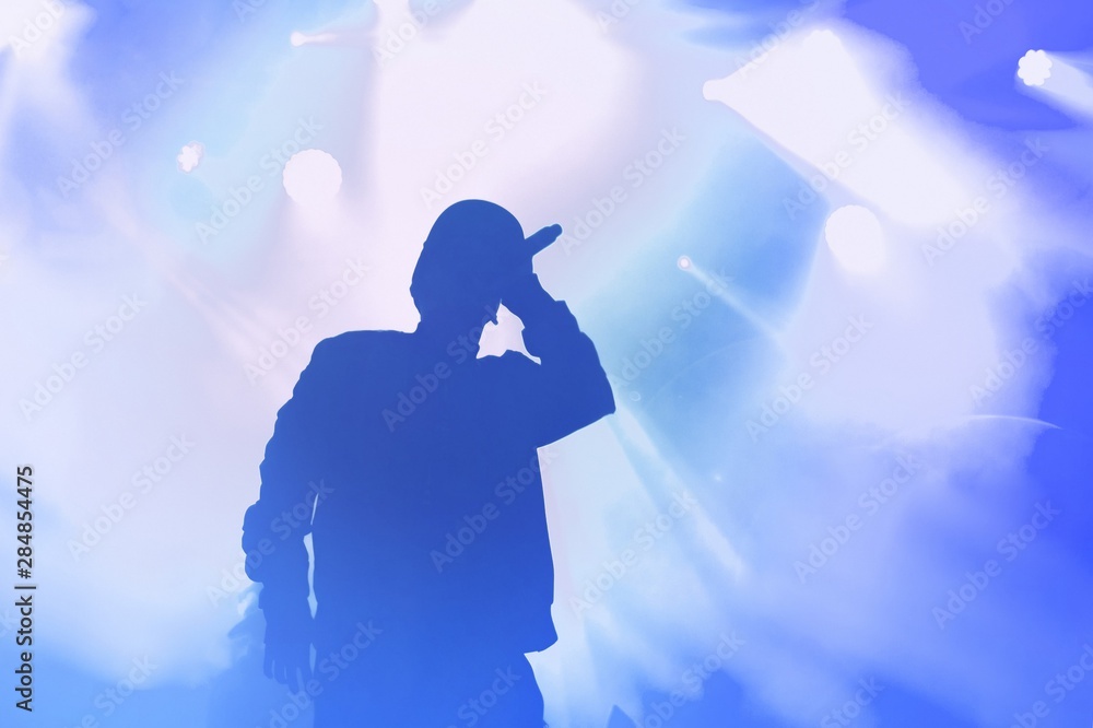 Rap singer with microphone on stage in music hall.Silhouette of rapper with mic in hand performing live on concert stage in nightclub.Rap artist with mic in bright blue stage lights singing song - obrazy, fototapety, plakaty 