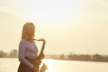 Wall Mural - silhouette of a young beautiful girl playing the saxophone at sunrise by the river, woman on the nature at sunrise giving a concert and relaxing, concept music