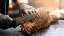 Grill Restaurant Kitchen. Chef In Black Cooking Gloves Using Knife To Cut Smoked Pork Ribs.