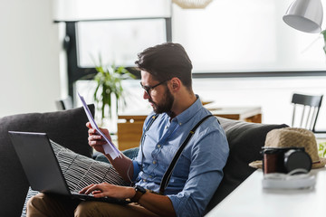 Young bearded businessman working on laptop computer while sitting on sofa at his home office. Business problems.