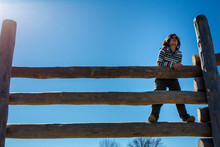 A Thoughtful Child Stands On A High Wooden Fence Against Blue Sky
