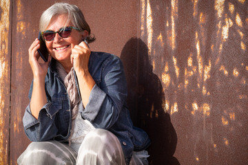 An attractive senior woman with gray hair talking with a cell phone leaning against a rusted iron panel. Sunset light. First cool days of autumn