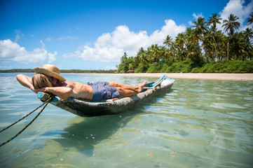 Tourist wearing a straw sun hat lying on a rustic dugout canoe off the shore of a sunny palm-lined tropical island beach in Bahia, Brazil