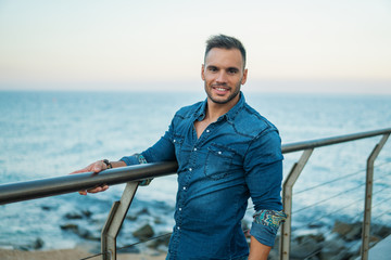 A young handsome man on the bridge near beautiful beach. Rich and successful male model wearing denim shirt