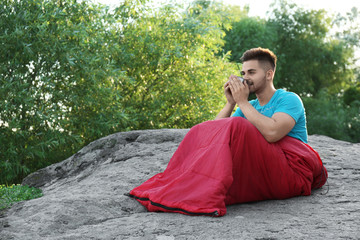 Sticker - Young man with cup in sleeping bag sitting on rock. Space for text