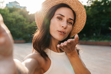 Image of pretty middle-aged woman taking selfie photo and sendng air kiss while walking in summer park