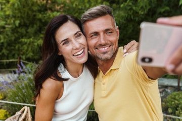 Wall Mural - Portrait of joyful middle-aged couple taking selfie photo on cellphone and hugging while walking in summer park