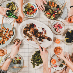 Wall Mural - Family or friends gathering dinner. Flat-lay of hands of people eating lamb shoulder, salads, vegetables, drinking wine over white checkered tablecloth, top view, square crop. Celebration party dinner