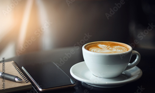 Photographie Closeup latte art coffee in white cup with smartphone and notebook on black table
