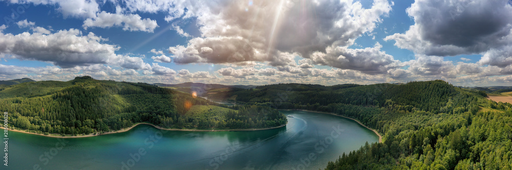 Camera flies over the Hennesee near Meschede and takes a wide view picture. You can see a ship, hills and forests, sunbeams and lens flares. Gorgeous nature in the Sauerland. - obrazy, fototapety, plakaty 