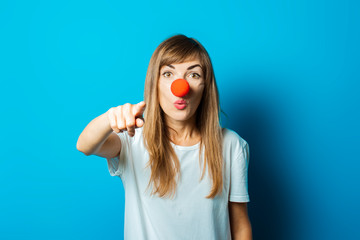 Beautiful young woman in a white t-shirt and a red clown nose with points her finger on a blue background. Concept party, costume, red nose day